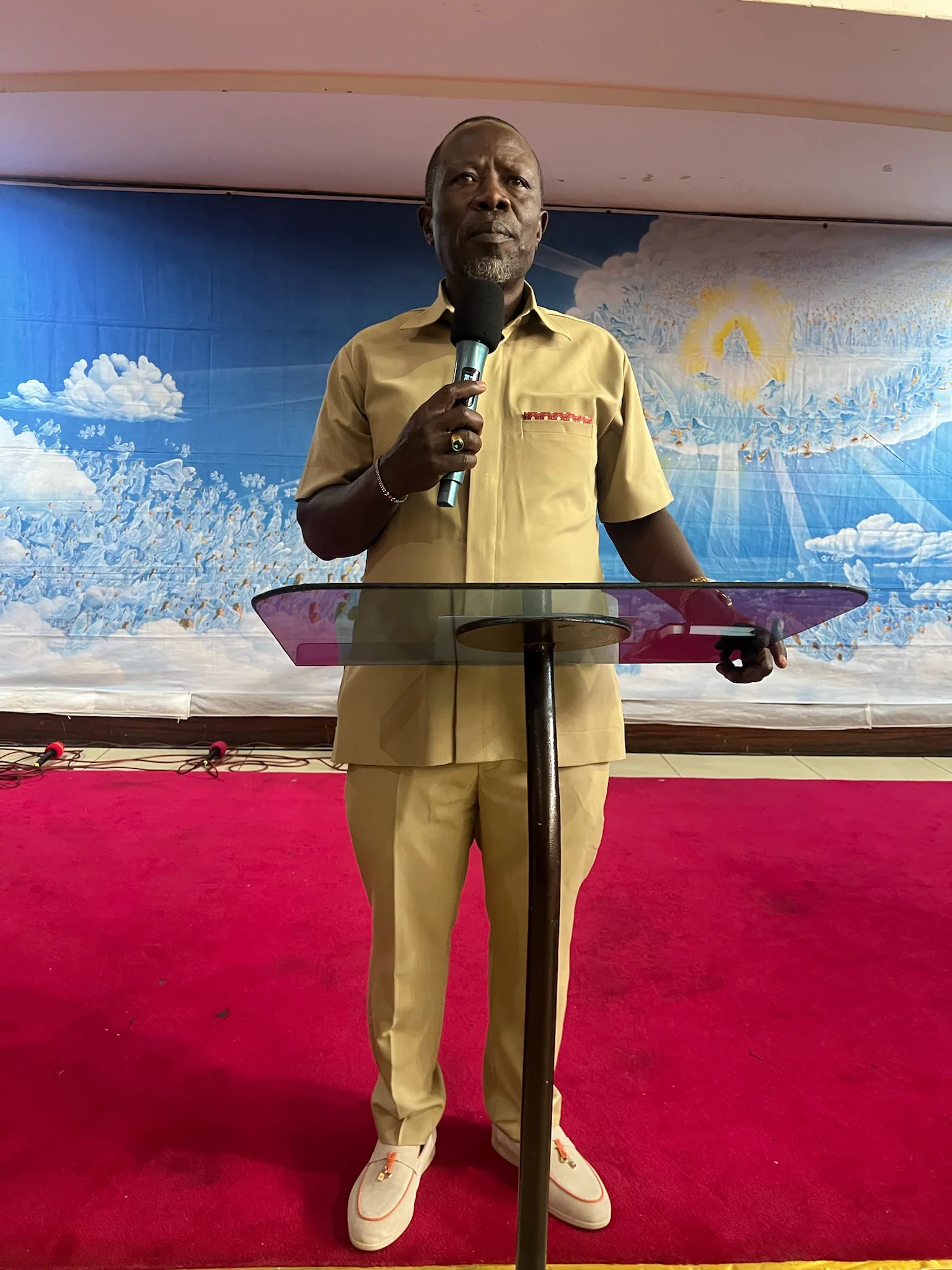 Pastor Adipo stands behind a clear pulpit holding a microphone as he addresses the gathering.