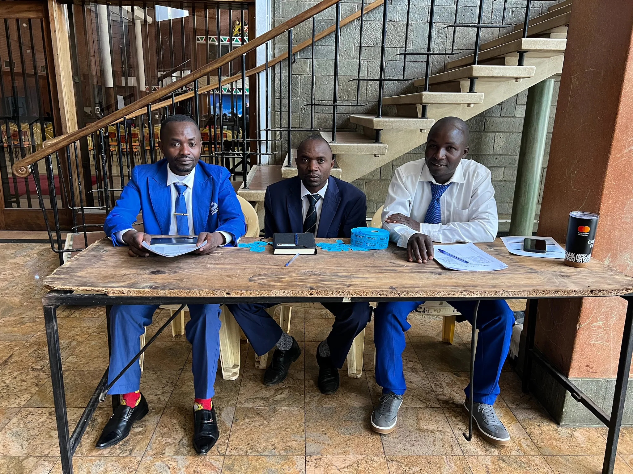 Three men sit behind a registration table with papers, a roll of blue raffle tickets, and event materials.