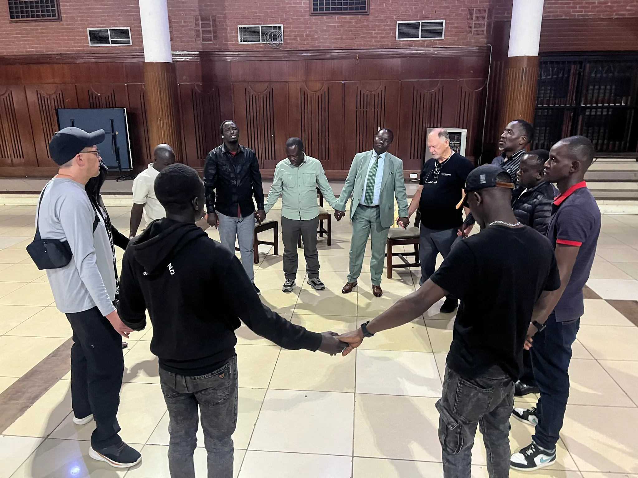 Michael Young, Don Kremer, Pastor Adipo, and local pastors stand in a circle holding hands in prayer inside Charter Hall.